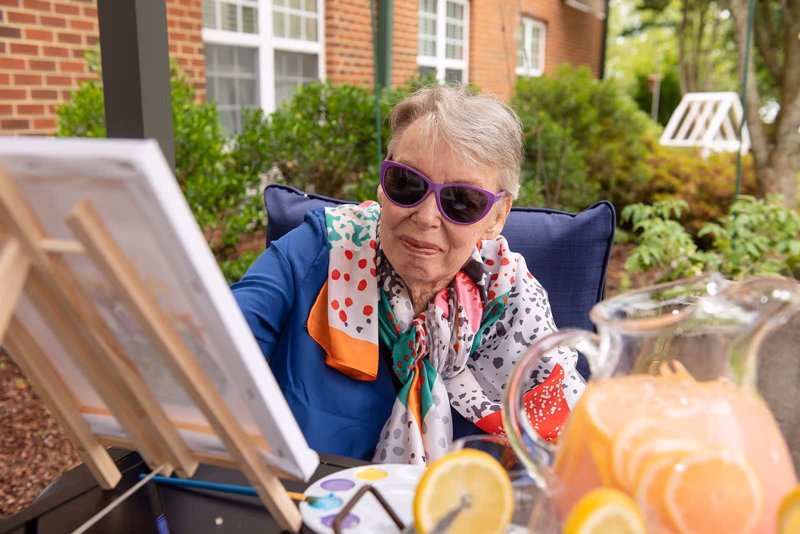 An elderly woman wearing purple sunglasses and a colorful scarf is sitting outdoors painting on a canvas set on an easel. In the foreground, there is a pitcher of lemonade with lemon slices and a glass of lemonade on the table. The background shows greenery and part of a brick building.
