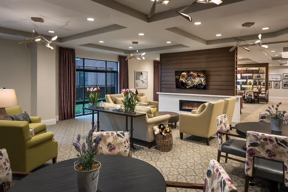 A cozy senior living facility common area with comfortable seating including green and beige armchairs and floral-patterned chairs around dark round tables. There is a modern electric fireplace with a TV mounted above it, showing a group of musicians playing French horns. The room has patterned carpet, ceiling lights, and large windows with dark curtains letting in natural light. Decorative plants and flowers are placed on tables, and a bookshelf with books and framed pictures is visible in the background.