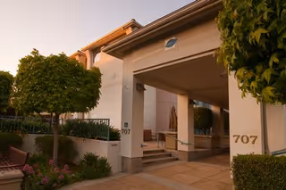 Entrance to a building at San Carlos Elms with the number 707 displayed on the wall. The entrance is covered by a roof supported by columns, with trees and shrubs surrounding the walkway leading up to the door.