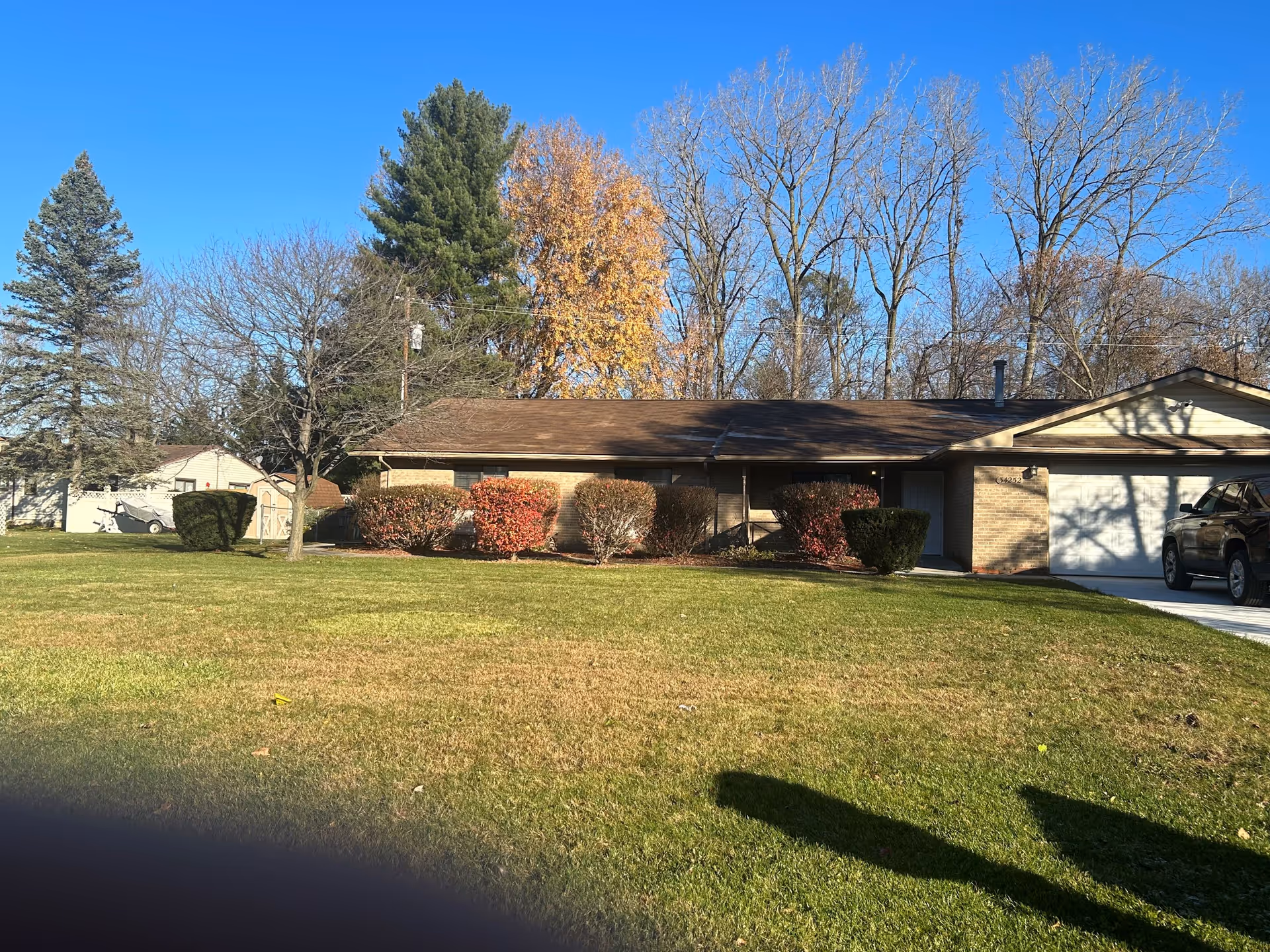 Single-story brick house with a brown roof, a white garage door, and a driveway with a black SUV parked. The front yard has green grass, trimmed bushes with some red foliage, and several trees with bare branches and some autumn leaves. The sky is clear and blue.