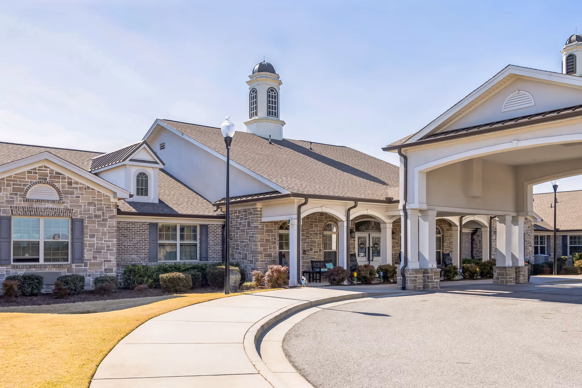 Exterior view of Barclay House of Aiken, showing a single-story building with stone and brick facade, a covered entrance with columns, a curved driveway, and a lamp post. The sky is clear and the landscaping includes bushes and grass.