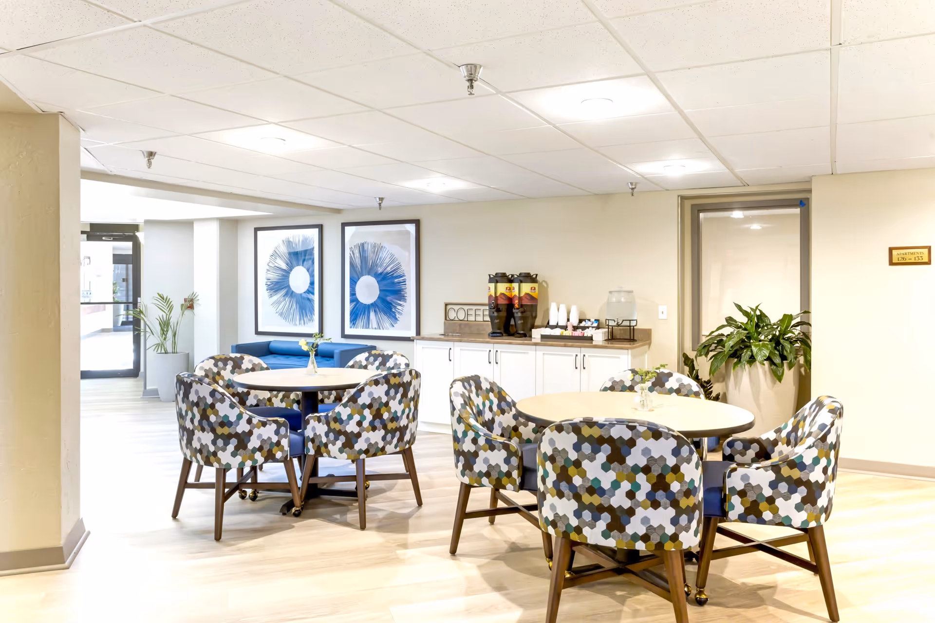 Bright communal dining area with round tables and patterned chairs, a beverage station, and wall art.