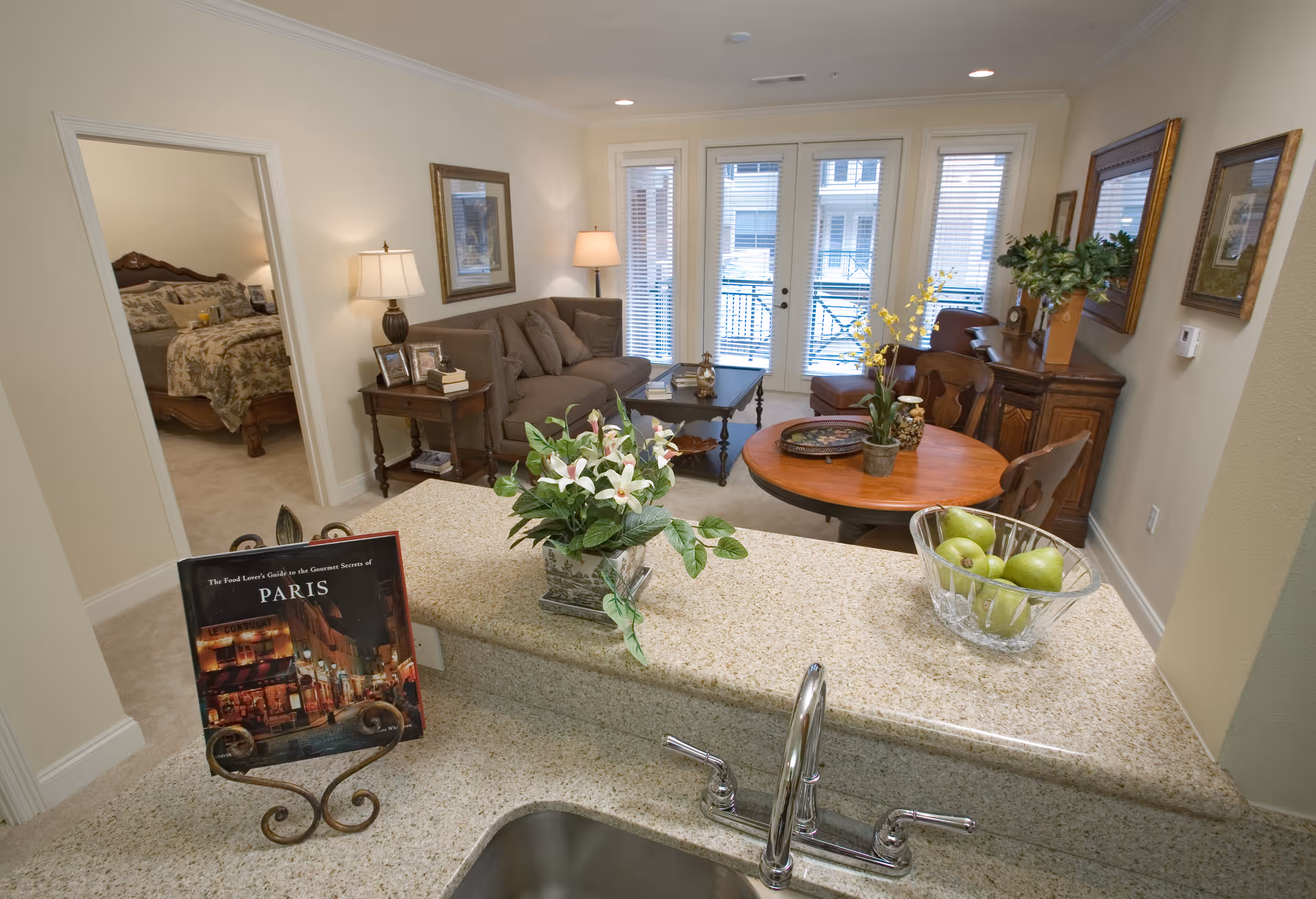 View from a kitchen sink area looking into a living room and dining area with a round wooden table, chairs, a sofa, and a bedroom visible through an open door. The living room has large windows with blinds and a door leading outside. The kitchen counter has a bowl of green apples and a vase with white flowers.