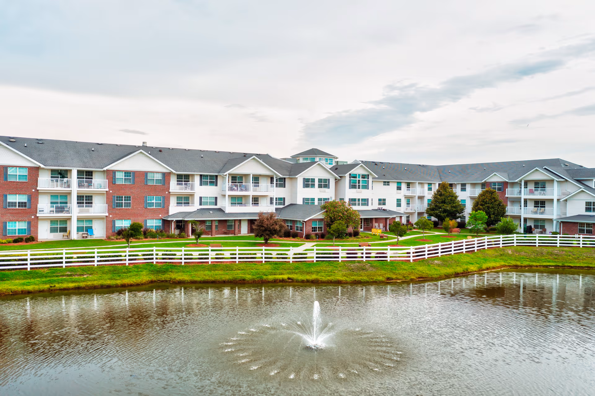 Exterior view of a large senior living facility building with multiple floors, balconies, and a white fence surrounding a landscaped area with trees and a pond featuring a water fountain in the foreground.
