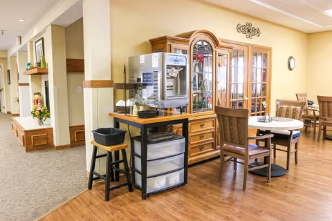 Common dining area with round tables and chairs, a coffee station on a small cart, and a glass-front cabinet against a beige wall.