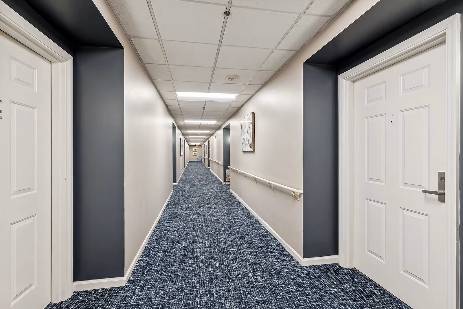 A long, well-lit hallway in The Lafayette facility with white walls, blue carpet, and white doors on either side. Handrails run along the right side of the hallway, and framed artwork is hung on the walls.