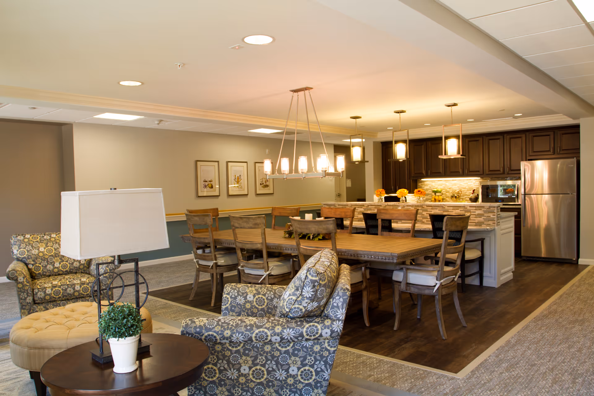 A spacious and well-lit common area in a senior living facility featuring a dining table with wooden chairs, a kitchen with dark wood cabinets, stainless steel refrigerator, and pendant lighting. In the foreground, there are patterned armchairs and a round wooden table with a lamp and a small potted plant.
