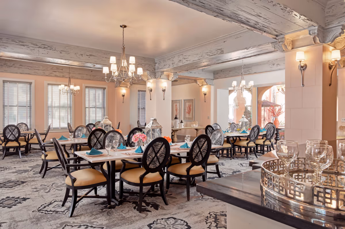 A bright and elegant dining room with multiple tables set for a meal. Each table has black chairs with beige cushions, teal napkins, and glassware. The room features large windows with blinds, chandeliers, wall sconces, and decorative beams on the ceiling. There are floral centerpieces on the tables and a decorative tray with glasses in the foreground.