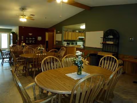 A dining hall with round wooden tables and chairs, and a kitchen area in back.