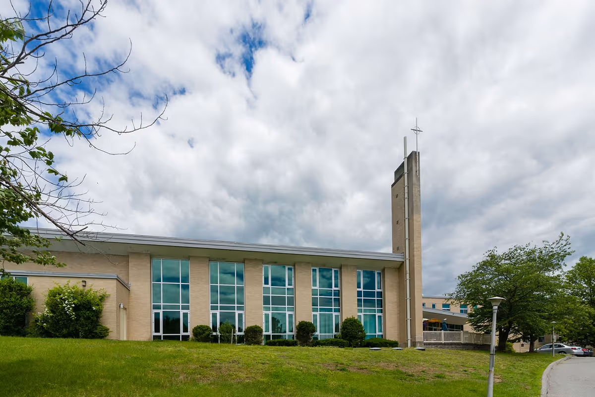 Exterior view of a beige brick building with large vertical windows and a tall tower with a cross on top. The building is surrounded by green grass, bushes, and trees under a cloudy sky.
