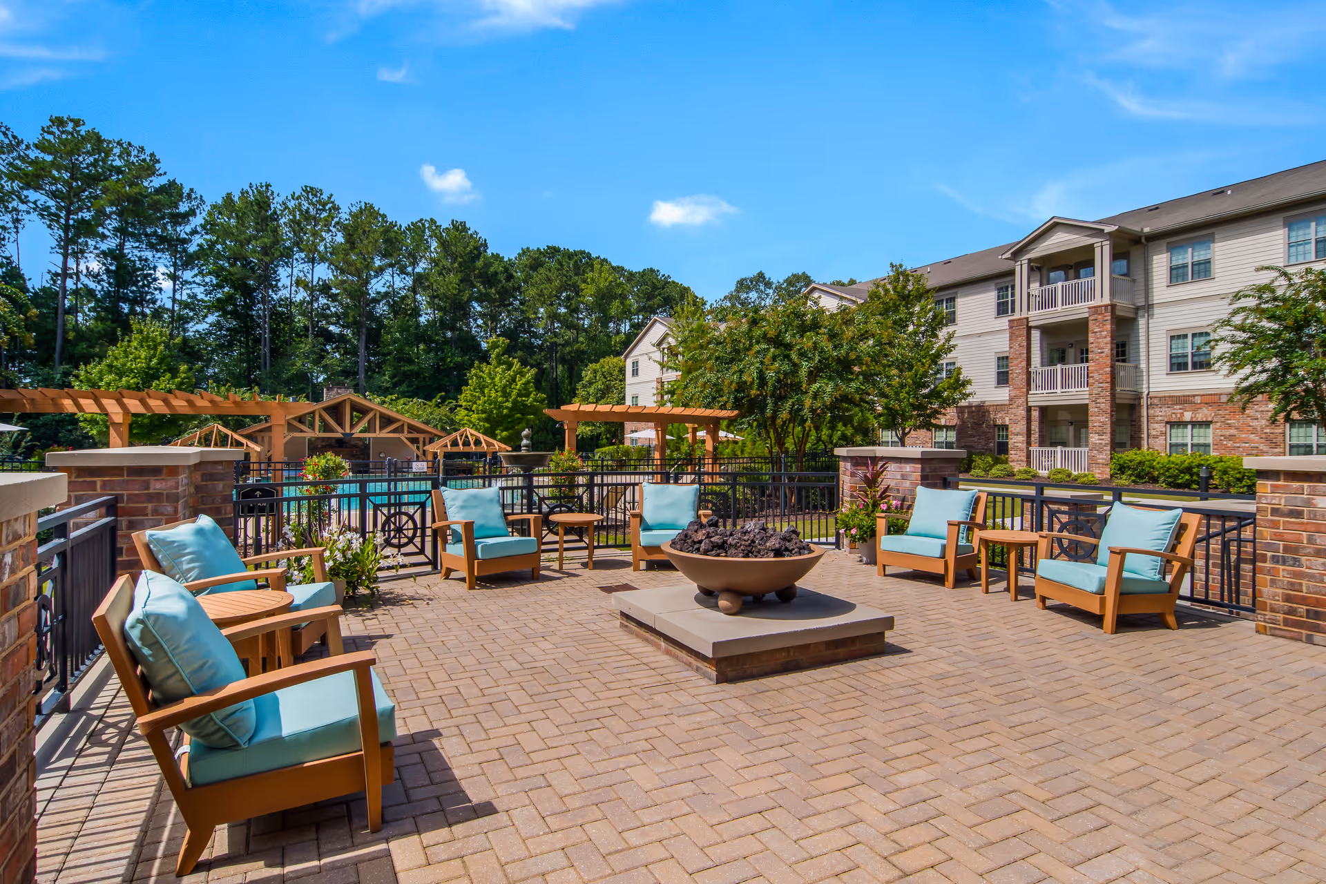 Outdoor patio area with cushioned wooden chairs arranged around a central fire pit. The patio is paved with bricks and surrounded by a black metal fence. In the background, there are trees, pergolas, and a multi-story residential building under a clear blue sky.