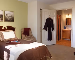 A cozy bedroom with a bed covered in brown and white bedding, a patterned armchair with a red throw blanket, and a door leading to a bathroom with a vanity, mirror, and shower curtain visible. A dark robe hangs on the door.