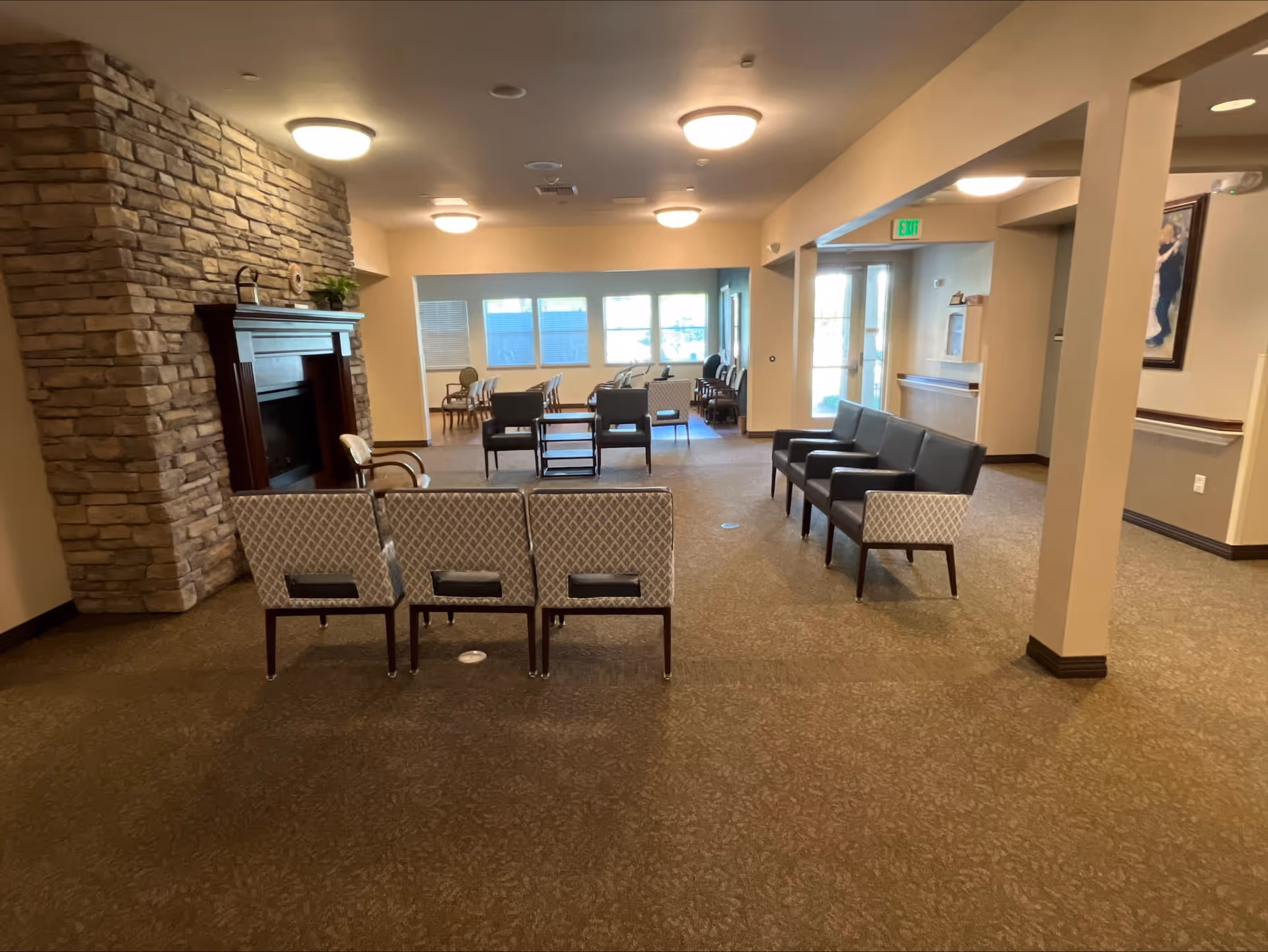 A spacious senior living facility common area with multiple seating arrangements including patterned chairs and black cushioned chairs. There is a stone fireplace on the left wall and large windows at the far end letting in natural light. The room has beige walls, carpeted floors, and ceiling lights. An exit sign is visible near a door on the right side.