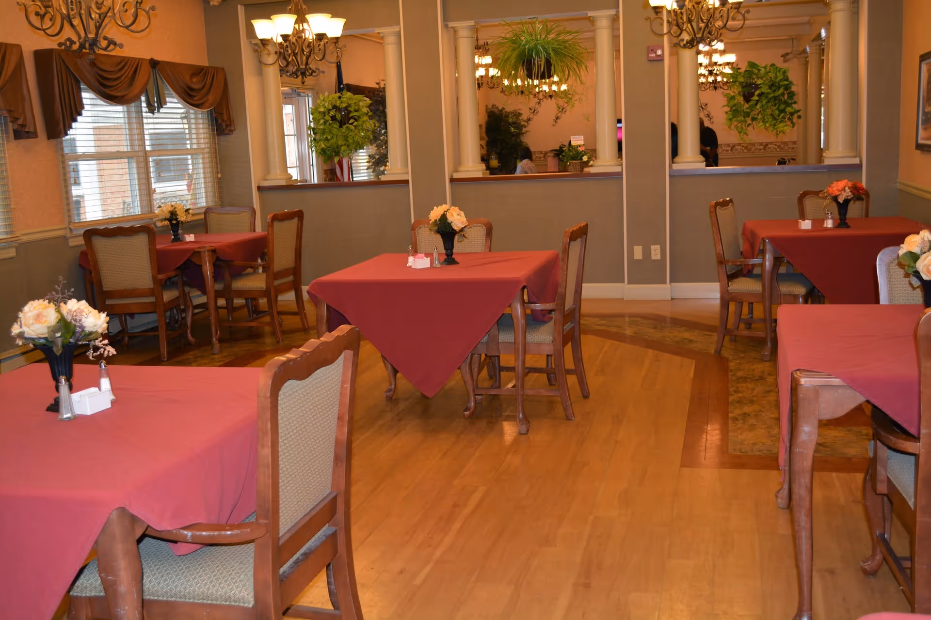 A dining room with several tables covered in red tablecloths, each with a small flower arrangement in a vase. Wooden chairs with cushioned seats surround the tables. The room has hardwood flooring, large windows with brown valances, and decorative chandeliers hanging from the ceiling. There are mirrors and plants on the far wall, creating a warm and inviting atmosphere.