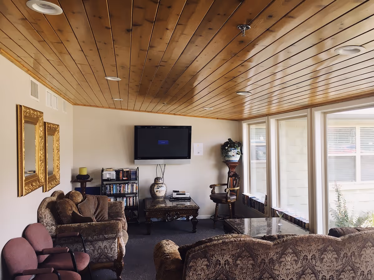 A cozy living room with patterned upholstered sofas and chairs, a wooden coffee table, a flat-screen TV mounted on the wall, a bookshelf with DVDs, two ornate gold-framed mirrors on the wall, and large windows letting in natural light. The ceiling is wooden with recessed lighting.