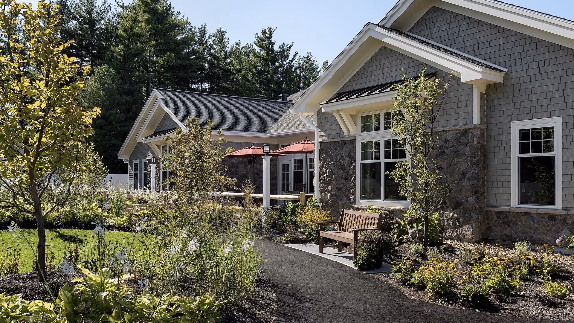 Outdoor view of a senior living facility with a paved walkway, landscaped garden beds with various plants and flowers, a wooden bench, and a building with gray siding and stone accents. There are red patio umbrellas and trees surrounding the area under a clear blue sky.