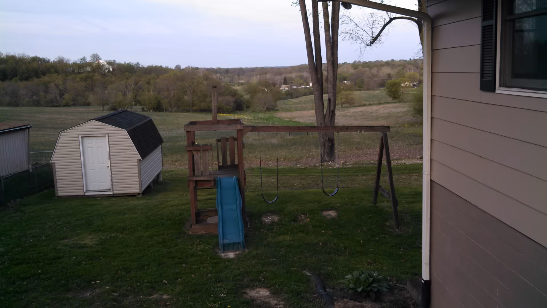 Backyard view showing a small wooden playset with a blue slide and two swings, a beige storage shed with a white door, green grass, and a rural landscape with trees and fields in the background. Part of a beige house with a window is visible on the right side.