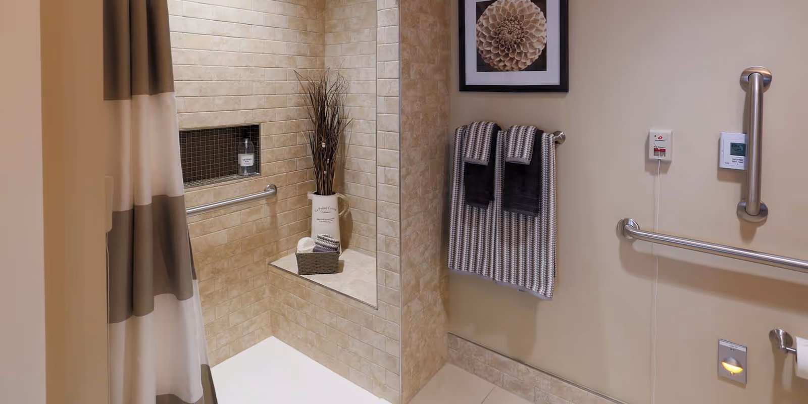 A beige tiled bathroom shower area with a built-in bench and a niche holding a bottle. A vase with decorative branches and a basket with towels are placed on the bench. On the adjacent wall, there are two striped towels hanging on a towel rack, a framed floral artwork, grab bars, a thermostat, and an emergency pull cord.