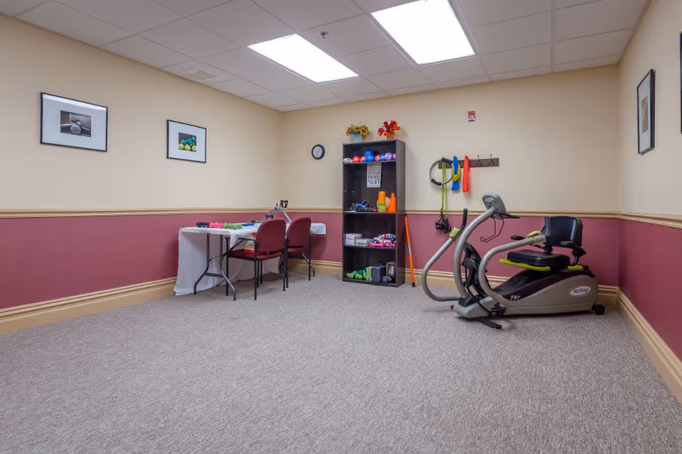 A small exercise room with beige and maroon walls, carpeted floor, and fluorescent ceiling lights. The room contains a recumbent exercise bike, a black shelving unit with various small exercise equipment like balls, cones, and weights, and a table with two red chairs. There are framed pictures on the walls and a clock above the shelving unit.