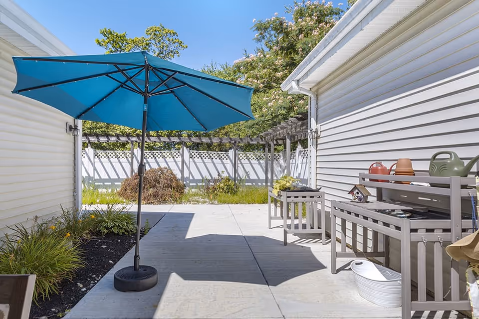 Outdoor patio area between two white buildings with a large blue umbrella providing shade. There are garden beds with plants on the left side and a wooden bench and table on the right side holding gardening tools and pots. A white fence and green trees are visible in the background under a clear blue sky.