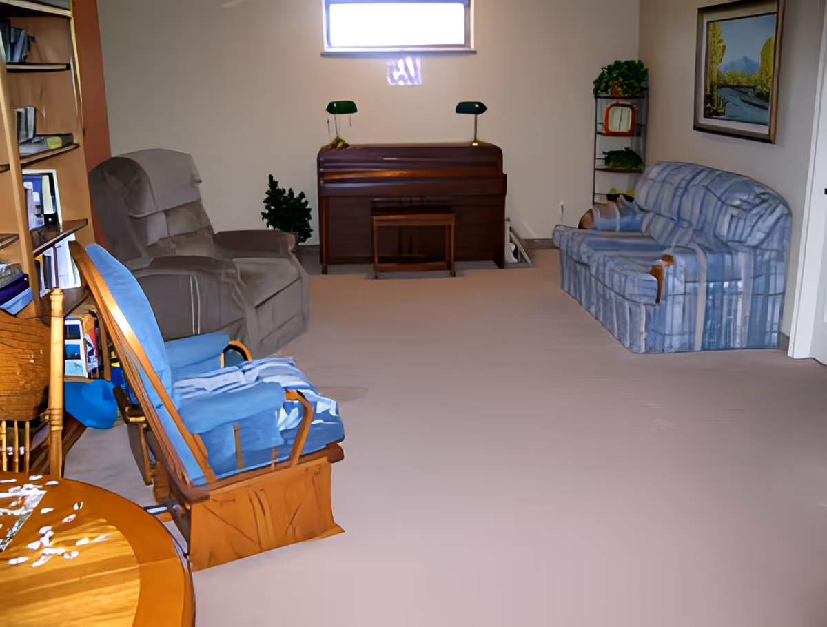 A cozy living room with a blue cushioned wooden rocking chair, a gray recliner, and a blue patterned sofa. There is a wooden piano against the far wall with two green lamps on top, a small wooden stool in front of it, and a small artificial plant beside it. A bookshelf filled with books and items is on the left side, and a framed landscape painting hangs on the right wall. The room has beige carpeting and a small window above the piano.