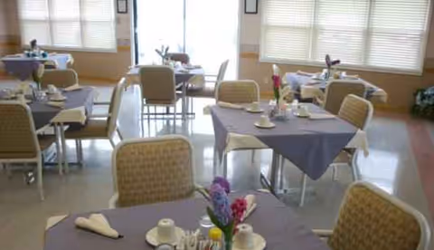 A bright dining room with purple tablecloths, arranged tables, and flower centerpieces.
