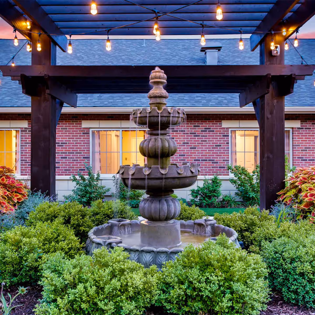A tiered stone water fountain surrounded by green bushes and colorful plants under a wooden pergola with string lights, with a brick building and lit windows in the background.