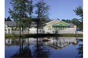 A single-story building with a green roof and white exterior is reflected in a calm pond in the foreground. Trees surround the building and the pond, creating a peaceful outdoor setting.