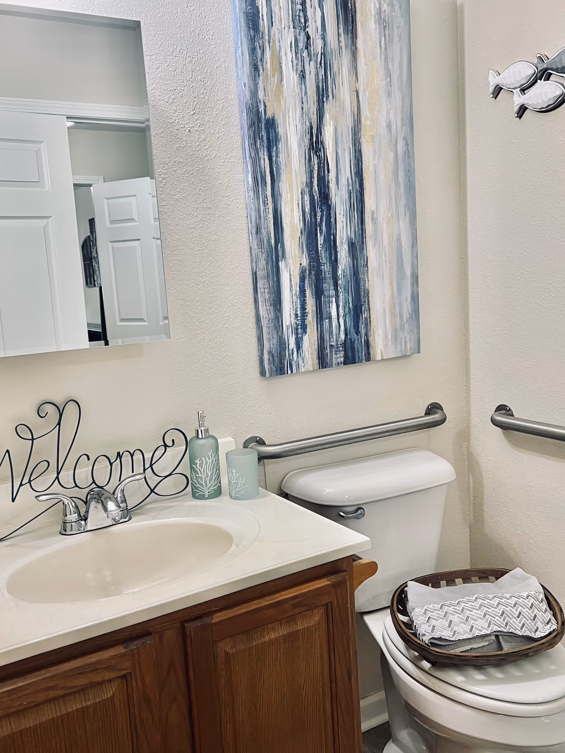 A bathroom with a white sink and wooden cabinet below, a silver faucet, and a mirror above the sink. On the countertop, there is a decorative 'Welcome' sign, a soap dispenser, and a tumbler with coral designs. A white toilet is next to the sink with a basket containing folded towels placed on the closed toilet lid. The walls are light-colored with a blue and white abstract painting hanging above the toilet and two silver grab bars installed on the walls.