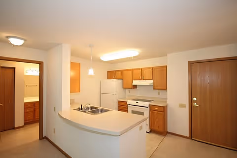 Open-plan apartment kitchen with a curved peninsula counter, sink, white refrigerator and stove, oak cabinets, and an entry door.