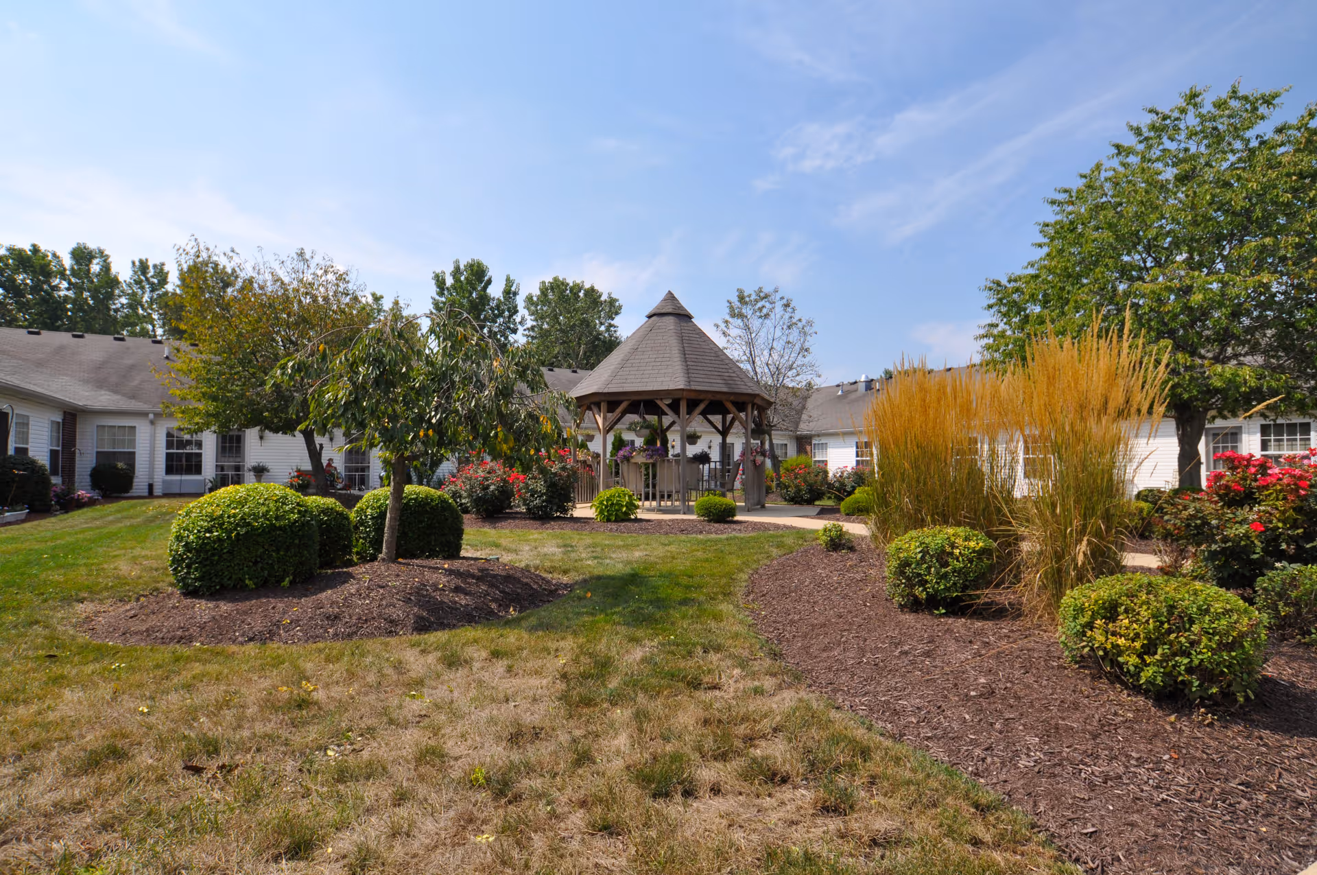 Outdoor garden area at Five Star Residences of Northwoods featuring a wooden gazebo surrounded by well-maintained bushes, trees, and flowering plants under a partly cloudy sky.