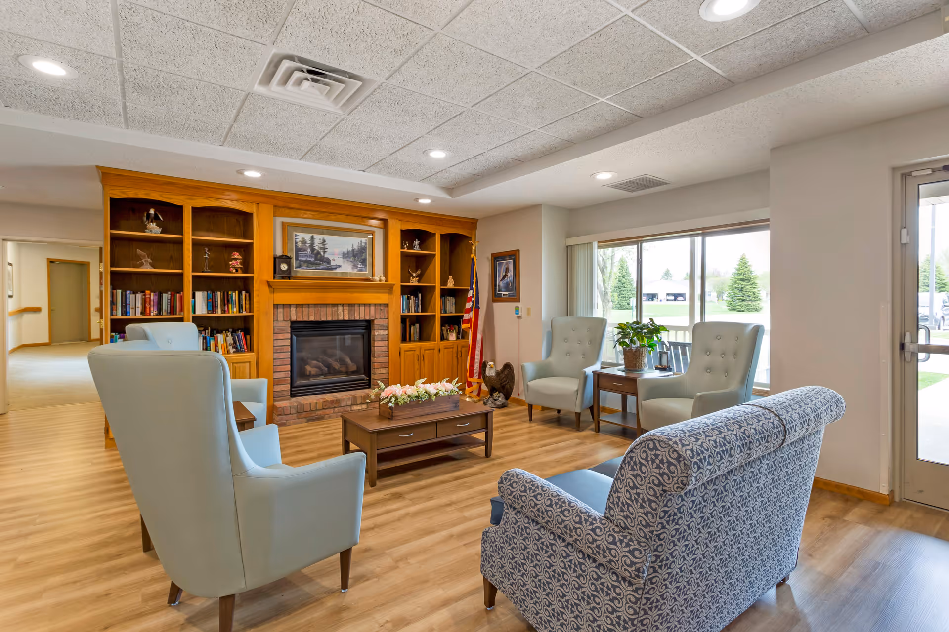 Bright senior living common room with armchairs and a patterned sofa arranged around a coffee table and a fireplace with built-in bookshelves.