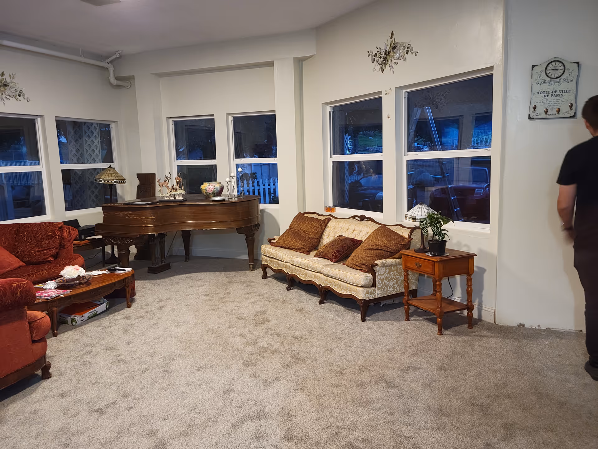 A cozy living room with a beige carpet, a vintage cream-colored sofa with brown cushions, a wooden side table with a small plant and lamp, a red upholstered armchair, a wooden coffee table with magazines and decorative items, and a grand piano near three windows. A person is partially visible on the right side of the image.