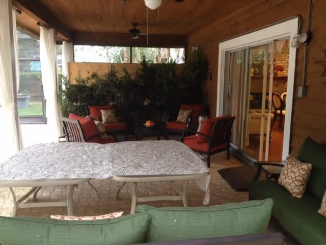 Covered outdoor patio area with cushioned seating including green and red chairs and sofas, a table covered with a white cloth, potted plants along the back, and sliding glass doors leading inside.