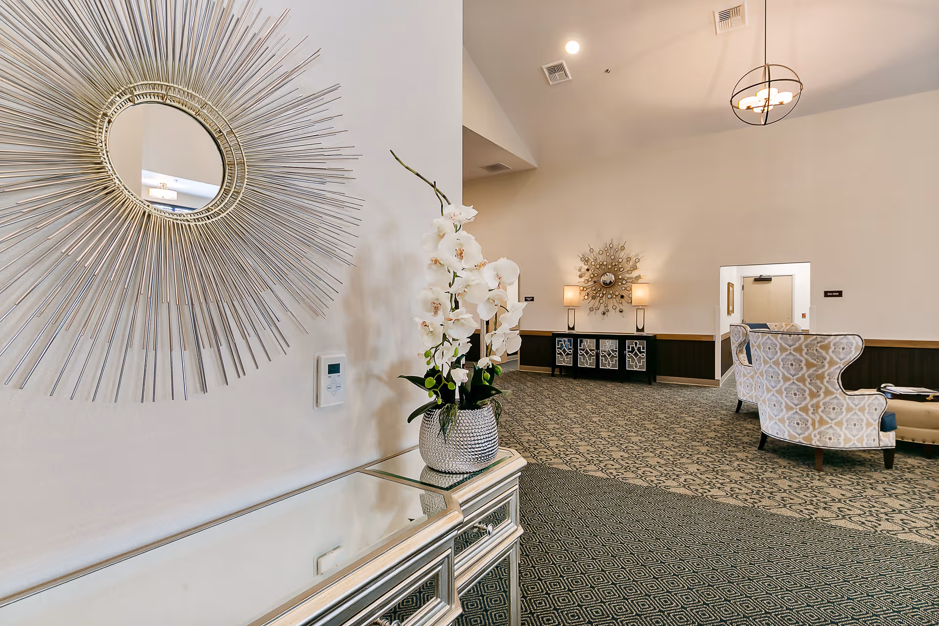 Interior view of a senior living facility lounge area featuring a decorative sunburst mirror on a white wall, a mirrored console table with a vase of white orchids, patterned carpet, two upholstered armchairs with a small table, and a sideboard with lamps and another sunburst mirror in the background.