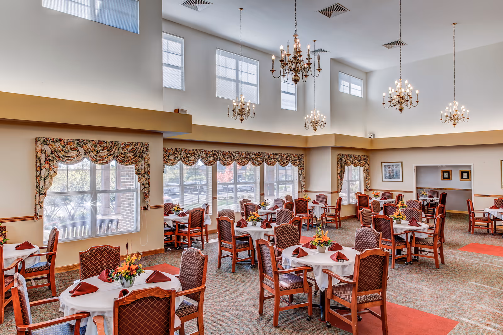 A spacious dining room with multiple round tables covered with white tablecloths, each set with folded burgundy napkins and floral centerpieces. The room features large windows with floral valances, high ceilings with several ornate chandeliers, and carpeted floors with red accent rugs. The walls are light-colored with wood trim, and there are framed pictures on the walls.