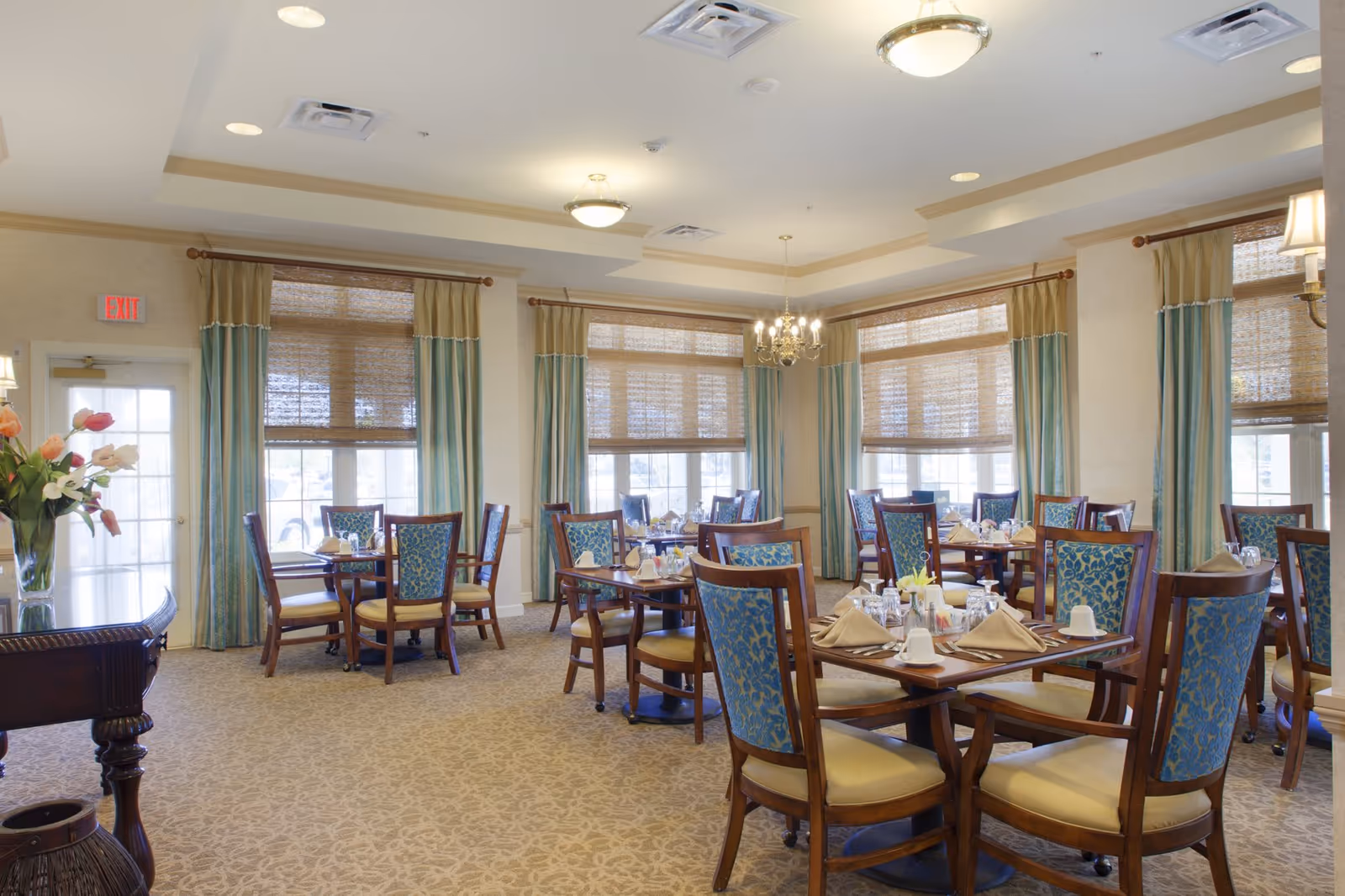 Dining room with round wooden tables set for meals and upholstered chairs beneath large windows with curtains.