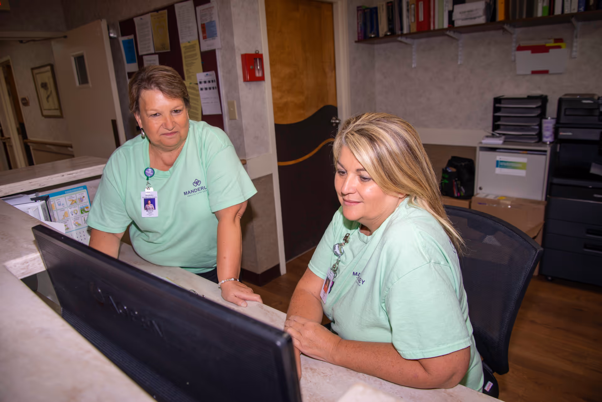 Two female staff members wearing light green Manderley Health Care Center shirts are working together at a reception desk, looking at a computer monitor in an office setting with shelves, files, and office equipment in the background.