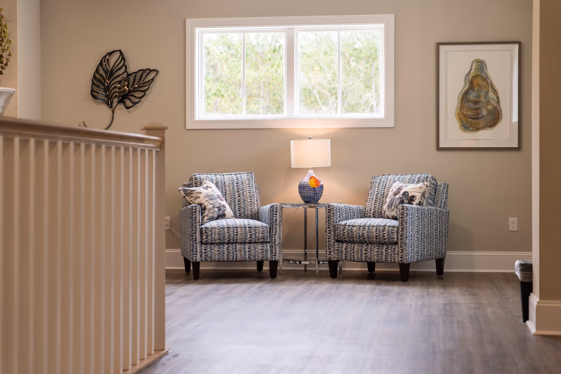 A cozy sitting area with two patterned armchairs separated by a small round table holding a blue lamp with a white shade. The wall behind features a large window with a view of greenery outside and a framed abstract artwork. The floor is wooden, and part of a white railing is visible on the left side.