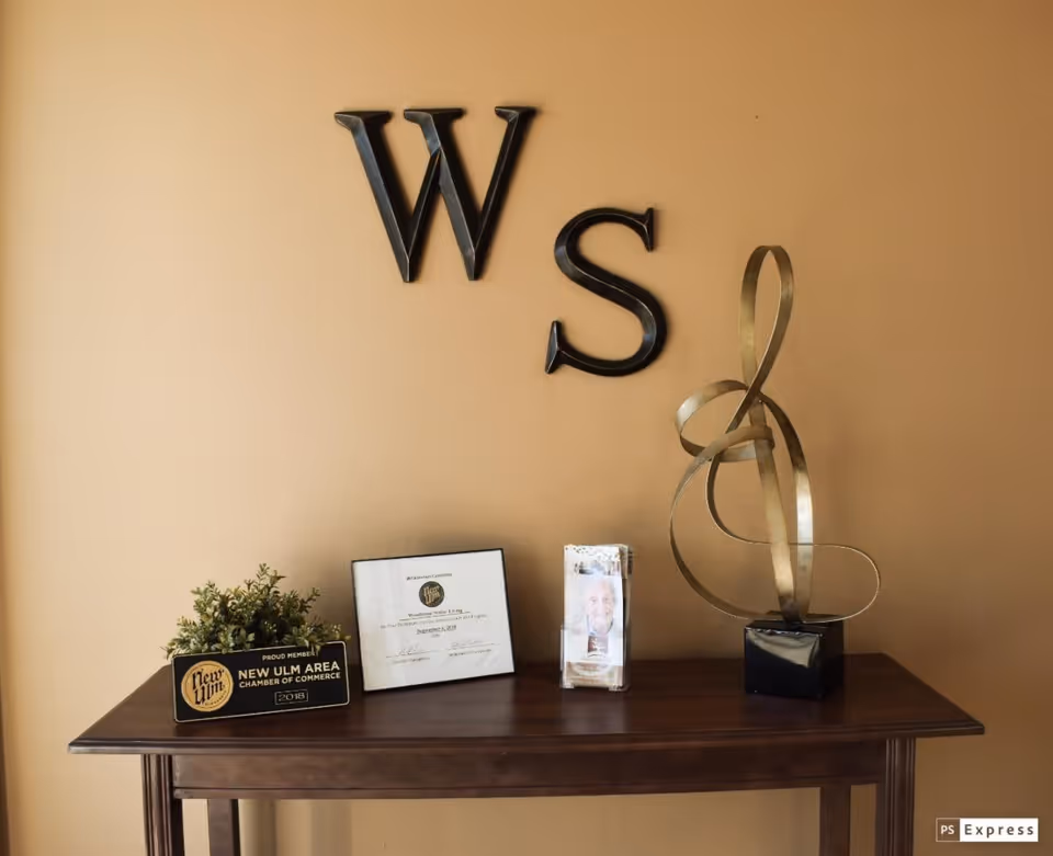 A wooden table against a beige wall with decorative metal letters 'W' and 'S' mounted above it. On the table are a small plant in a New Ulm Area Chamber of Commerce plaque, a framed certificate, a brochure holder with a smiling elderly person's photo, and a modern abstract metal sculpture.