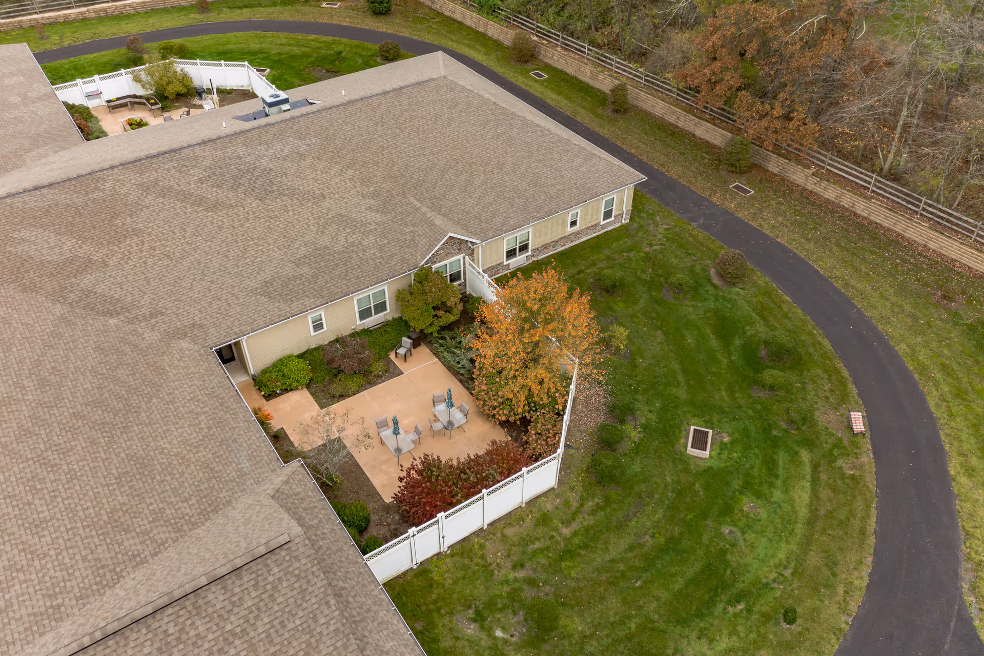 Aerial view of a senior living facility courtyard with a fenced patio area containing outdoor chairs and tables surrounded by trees and shrubs. The building has a beige exterior and a shingled roof. A curved paved pathway runs alongside the building with green grass and some trees nearby.