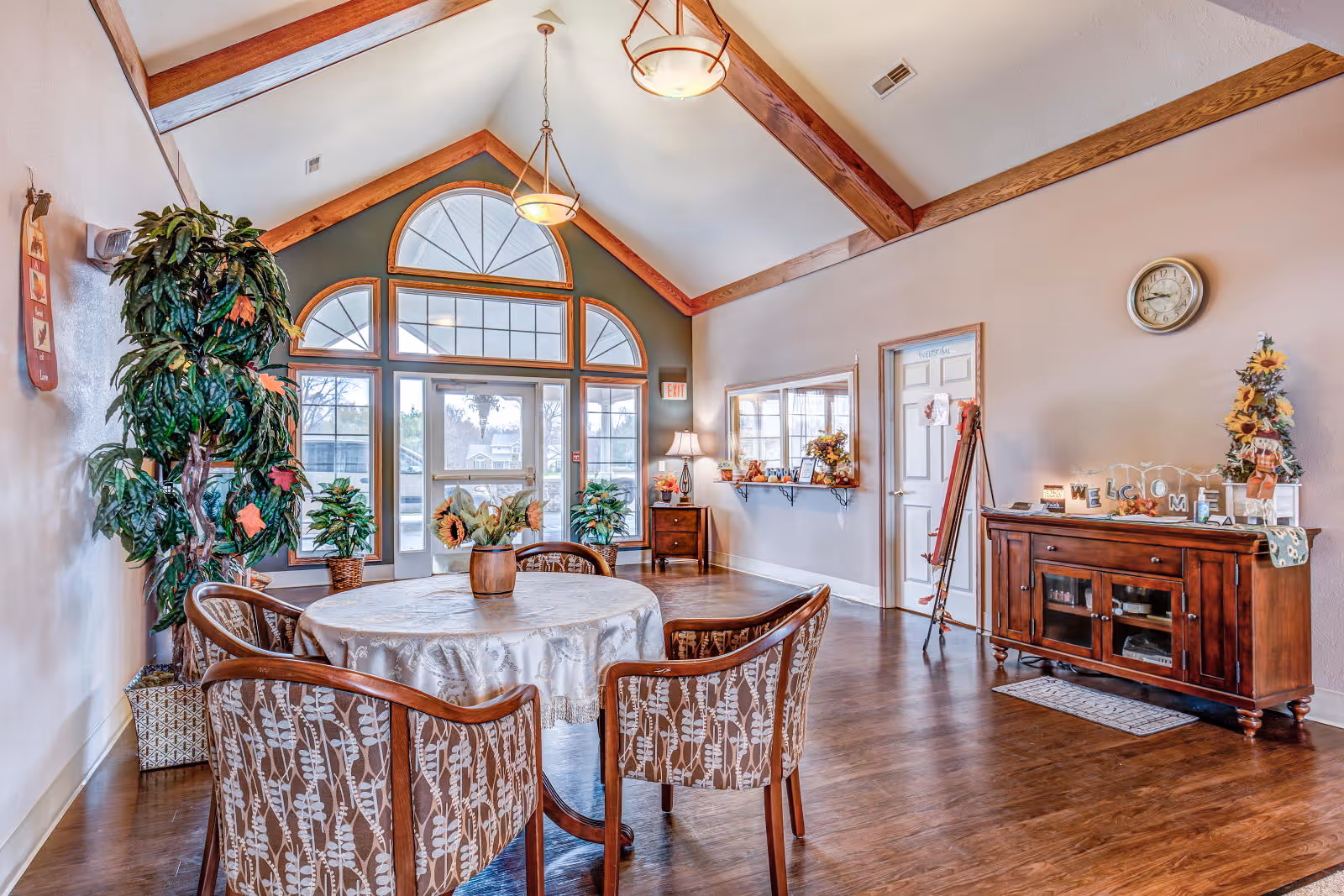 Communal dining area with a round table and four patterned chairs, large arched windows, exposed wood beams, plants, and a wooden sideboard.