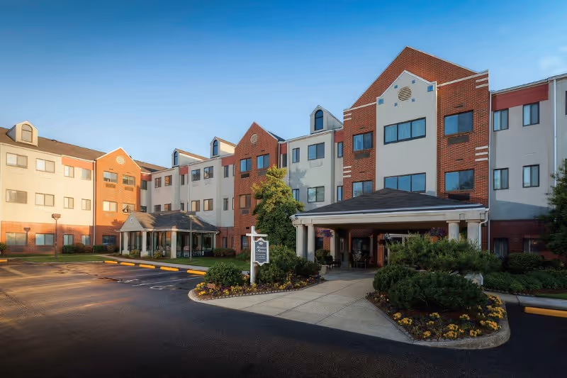 Front entrance and facade of a multi-story brick-and-stucco retirement community with a covered drop-off and landscaped driveway.