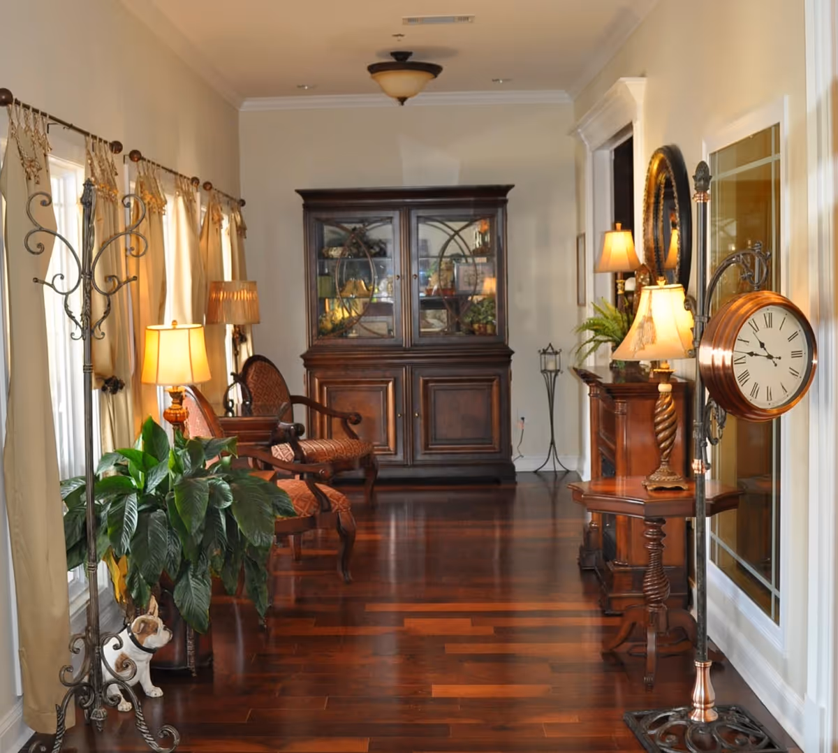 A warmly lit interior hallway with polished wooden floors, featuring a large wooden cabinet with glass doors at the end, upholstered chairs along the left wall, several table lamps, a standing clock with Roman numerals on the right, and a coat rack with a small dog statue at the base near the windows with beige curtains.