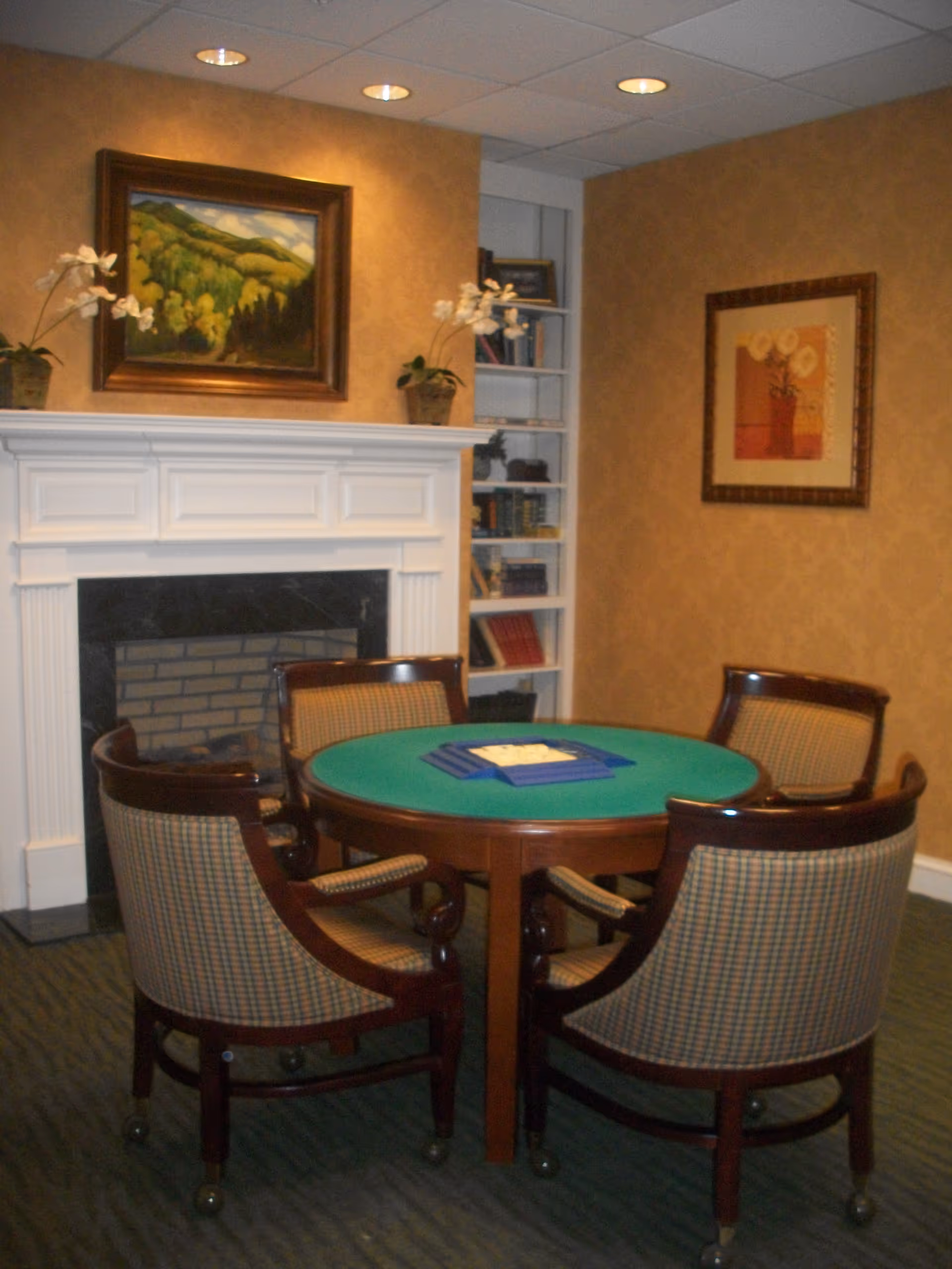 A cozy room with a round green felt card table surrounded by four cushioned wooden chairs on casters. Behind the table is a white fireplace with a framed landscape painting above it. On either side of the fireplace are potted plants with white flowers. To the right, there is a built-in bookshelf with books and decorative items. The walls are painted a warm beige color and have another framed artwork hanging.