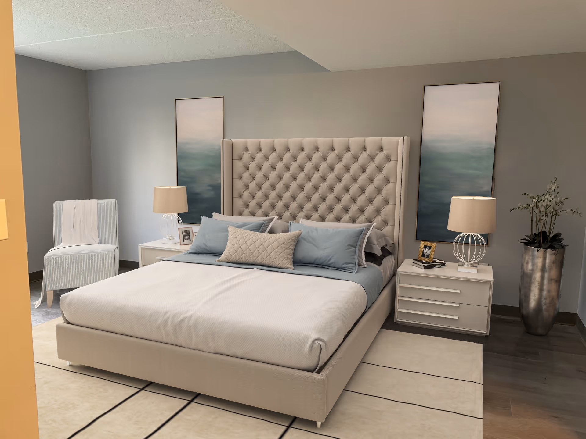 A neatly made bed with a tufted beige headboard in a bedroom. The bed has light blue and beige pillows and a beige bedspread. On either side of the bed are white nightstands with modern lamps, framed photos, and books. Two tall abstract paintings hang on the gray wall behind the bed. A light blue chair with a white throw is positioned in the corner, and a tall silver vase with white flowers stands to the right. The floor has a light-colored rug with dark lines and dark wood flooring.