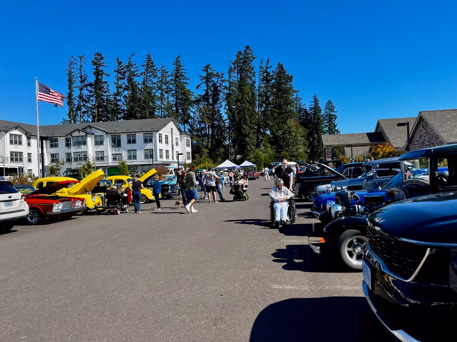 Outdoor car show event in a parking lot with vintage and classic cars lined up on both sides. People, including some in wheelchairs, are walking and observing the cars. A large white building and tall trees are visible in the background under a clear blue sky. An American flag is flying on a flagpole near the building.