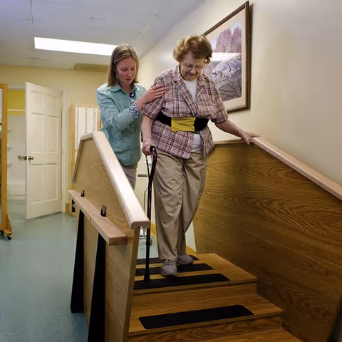 An elderly woman using a cane is assisted by a caregiver as she carefully walks down a short set of wooden stairs with handrails on both sides inside a rehabilitation facility.