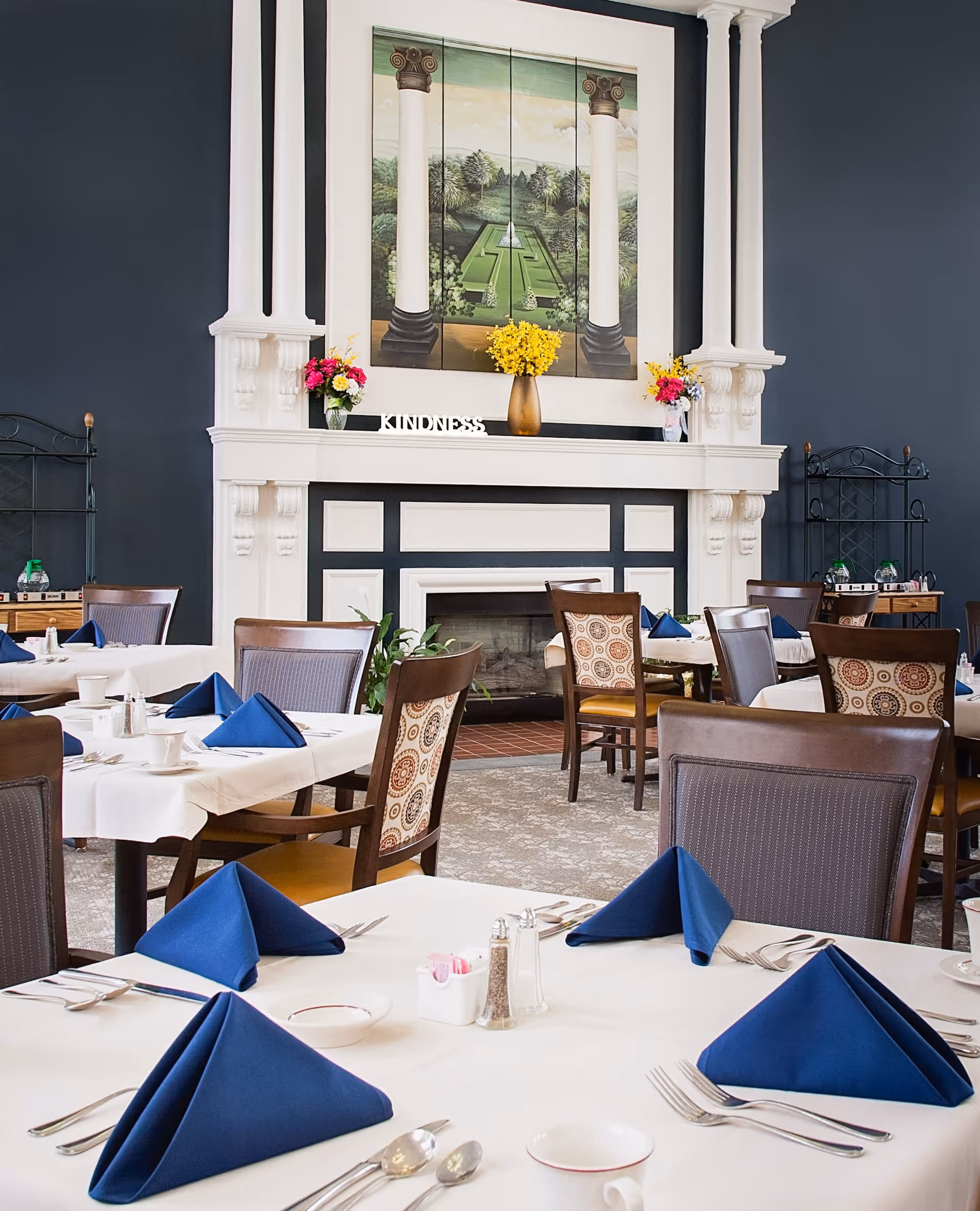 Dining room with tables covered in white tablecloths and set with blue folded napkins, silverware, and cups. The room features a decorative white fireplace with a painting above it, flanked by two flower arrangements and the word KINDNESS displayed on the mantel.
