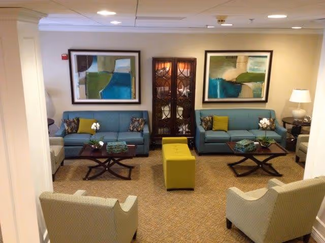 Communal living room with two blue sofas, two armchairs, coffee tables, framed abstract art, and a green ottoman.